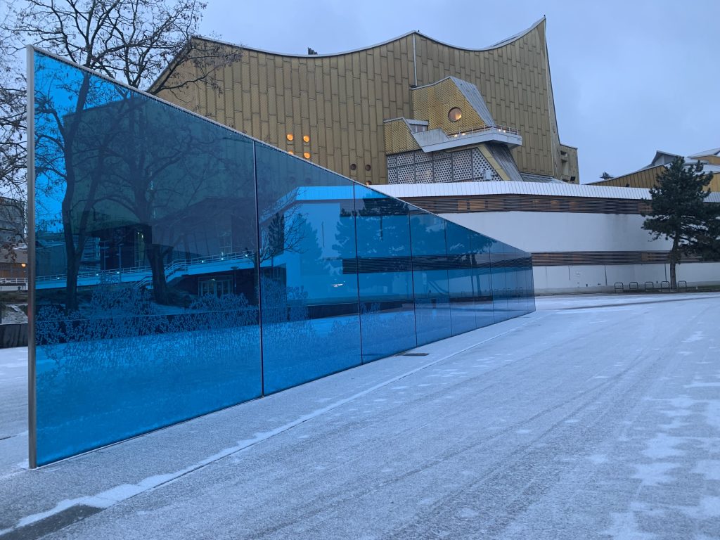 Gedenk- und Informationsort für die Opfer der NS- „Euthanasie“-Morde im Winter. Eine blaue Glaswand steht im Schnee, im Hintergrund ist die Philharmonie.