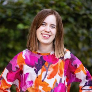 Porträt einer Person mit schulterlangen, braunen Haaren und einem bunt gemusterten Oberteil. Im Hintergrund ist eine grüne Pflanze.