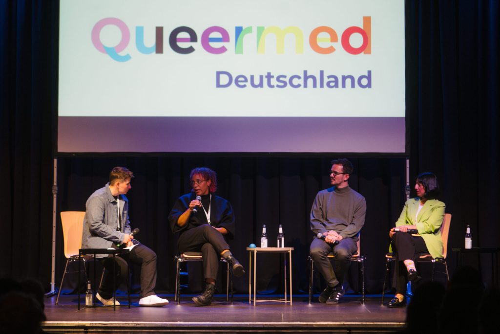 4 Personen sitzen auf einer Bühne und sind im Gespräch, darüber ein Banner „Queermed Deutschland“ in Regenbogenfarben.