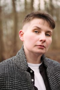 Weiß gelesene Person mit Untercut und kurzen, braunen Haaren blickt aufmerksam in die Kamera. Sie hat blaue Augen, trägt einen silbernen Ring in der Mitte des Ohrs, einen grau-schwarzen Blazer, darunter ein schwarzes, offenes Hemd und darunter ein weißes Shirt.