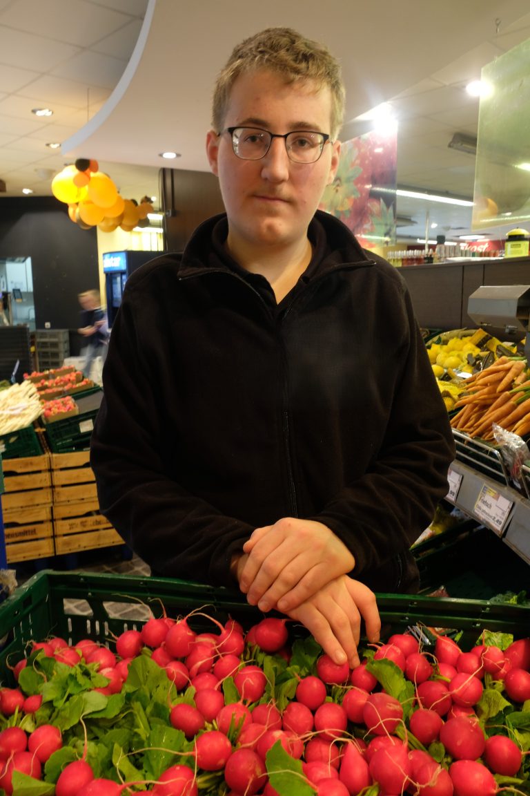 Person mit kurzen Haaren und Brille steht in schwarzem Hemd vor einer Kiste mit Radischen.