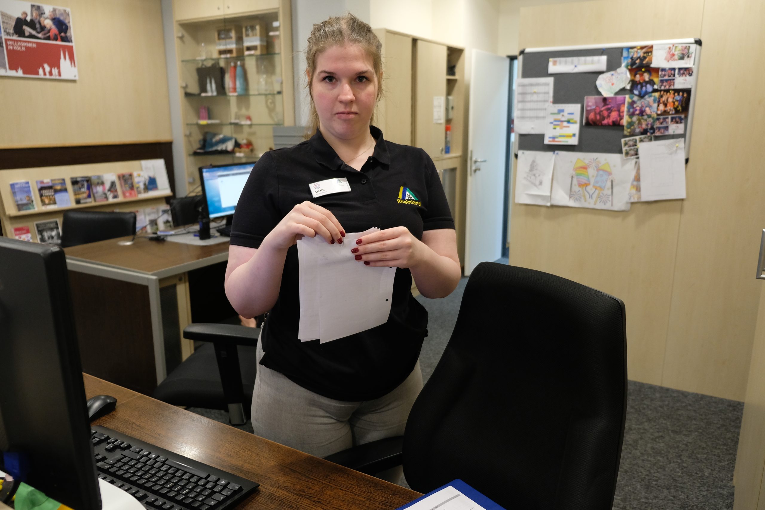 Person mit blondem Zopf steht in Arbeitskleidung hinter einem Empfangstresen und hält ein Blatt Papier in den Händen.