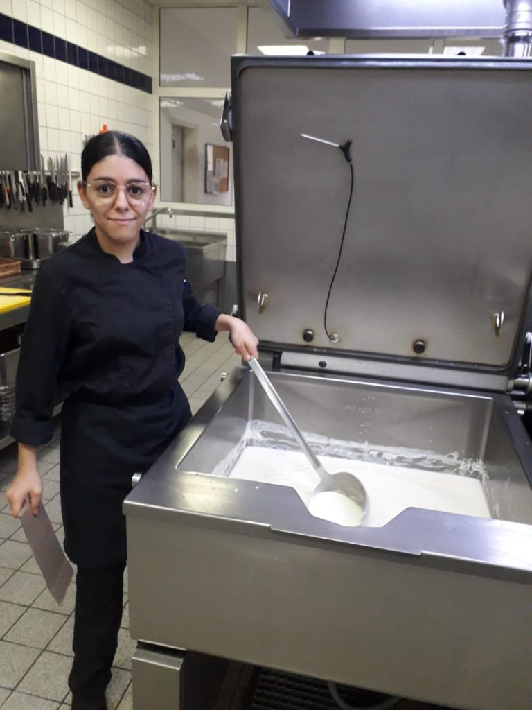Eine Person mit zusammengebundenen, schwarzen Haaren und schwarzer Gastronomiebekleidung in einer Küche mit einem großen Kochlöffel in der Hand, in der eine weiße Flüssigkeit ist.