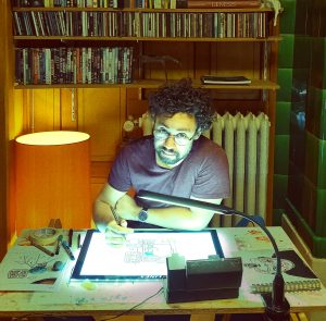 Person mit Brille, dunklen Locken und Bart sitzt an einem Schreibtisch und arbeitet an einem digitalen Zeichenbrett. Im Hintergrund sind DVDs und CDs im Holzregal.