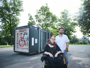 Jasmin Polsini und Valentin Weilenmann der Initiative „Schlafende Hunde wecken“ sind vor ihrer Kunstinstallation zu sehen. Sie sitzt in einem Aktivrollstuhl und trägt schwarze Kleidung, Er steht in weißem Shirt und Jeans daneben.