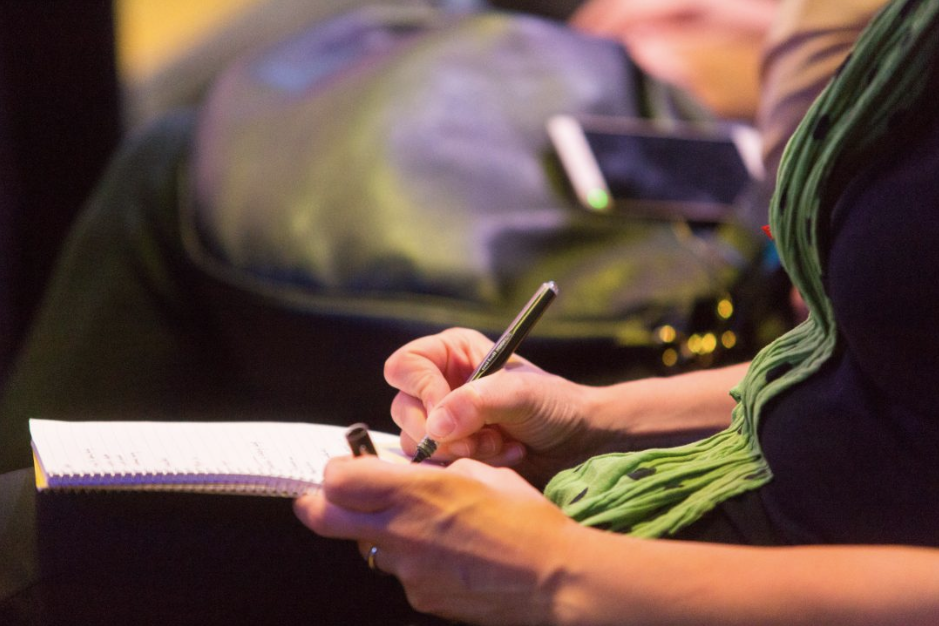 Ausschnitt einer weiß positionierten Person, von der man beide Arme und Hände sieht, sie Schreibt etwas auf einen Zettel. Die Person trägt einen grünen Schal, im Hintergrund ist eine Tasche und ein Handy zu sehen.