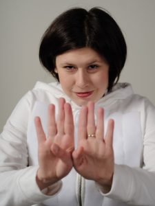 Eine weiß gelesene Person im weißen Hoodie streckt beide Handflächen nach vorne, die Daumen aneinander. Sie trägt einen Ring am linken Mittelfinger. Ihre dunklen Haare sind schulterlang und offen, sie trägt roten Lippenstift und lächelt leicht.