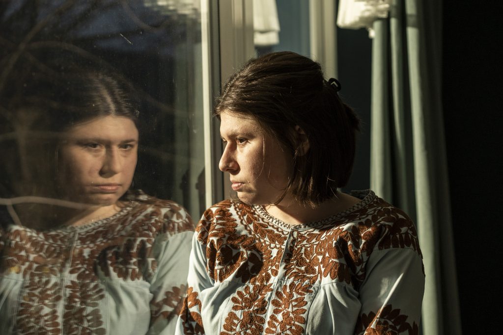 Eine Frau mit kurzen braunen Haaren steht am Fenster und schaut nachdenklich nach draußen; ihr Gesicht spiegelt sich im Glas. Warmes Licht fällt seitlich auf ihr Gesicht und hebt die ernste, ruhige Stimmung hervor.