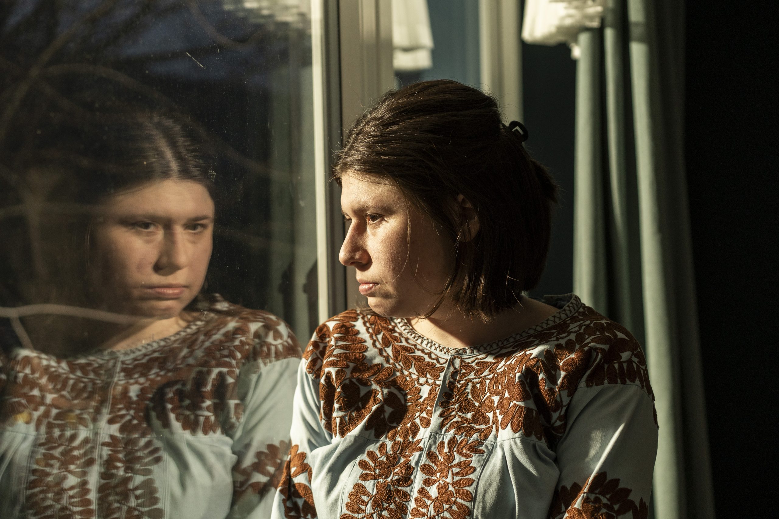 Eine Frau mit kurzen braunen Haaren steht am Fenster und schaut nachdenklich nach draußen; ihr Gesicht spiegelt sich im Glas. Warmes Licht fällt seitlich auf ihr Gesicht und hebt die ernste, ruhige Stimmung hervor.