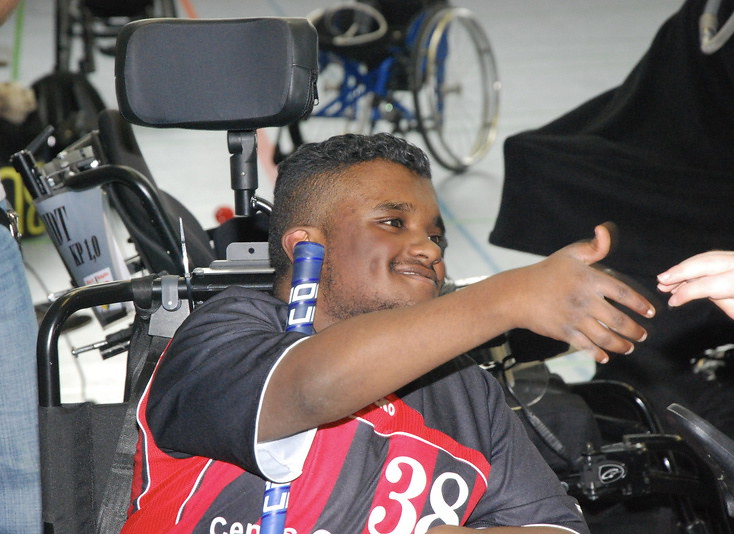 Schwarz positionierte Person im E-Rolli und Trikot reicht einer Person die Hand und lächelt.