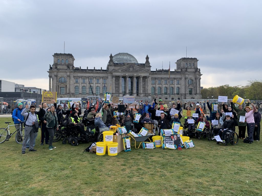 Mehrere Menschen mit Behinderung demonstrieren vor dem Deutschen Bundestag. Sie halten bunte Schilder in den Händen und strecken die linke Faust nach oben.