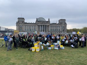 Mehrere Menschen mit Behinderung demonstrieren vor dem Deutschen Bundestag. Sie halten bunte Schilder in den Händen und strecken die linke Faust nach oben.