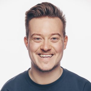 Porträt einer weiß positionierten Person mit kurzen, hellbraunen Haaren und blauem Shirt, die in die Kamera lacht.