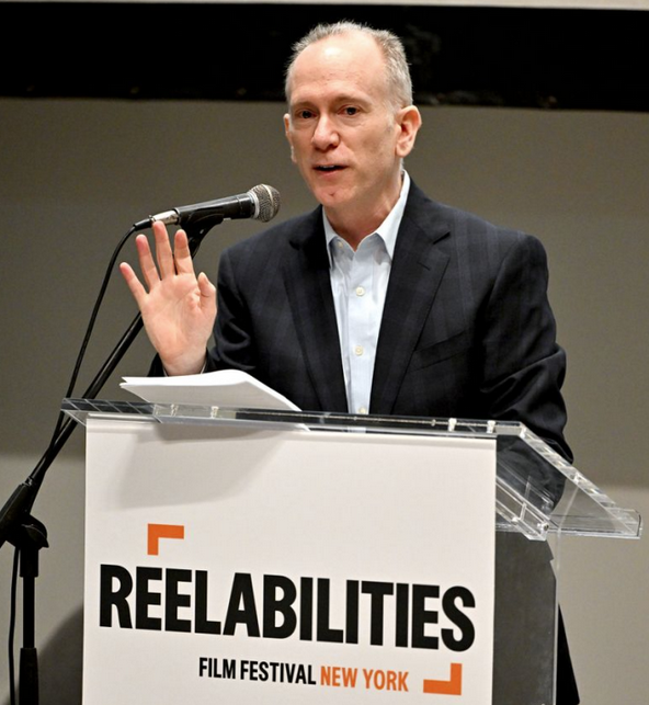 Weiß positionierte Person im Anzug steht an einem Pult mit der Aufschrift „ReelAbilities“.