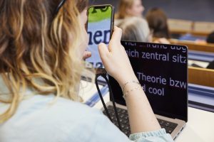 Weiß positionierte Person sitzt in Hörsaal und nutzt ein Screenreader mit Vergrößerung.