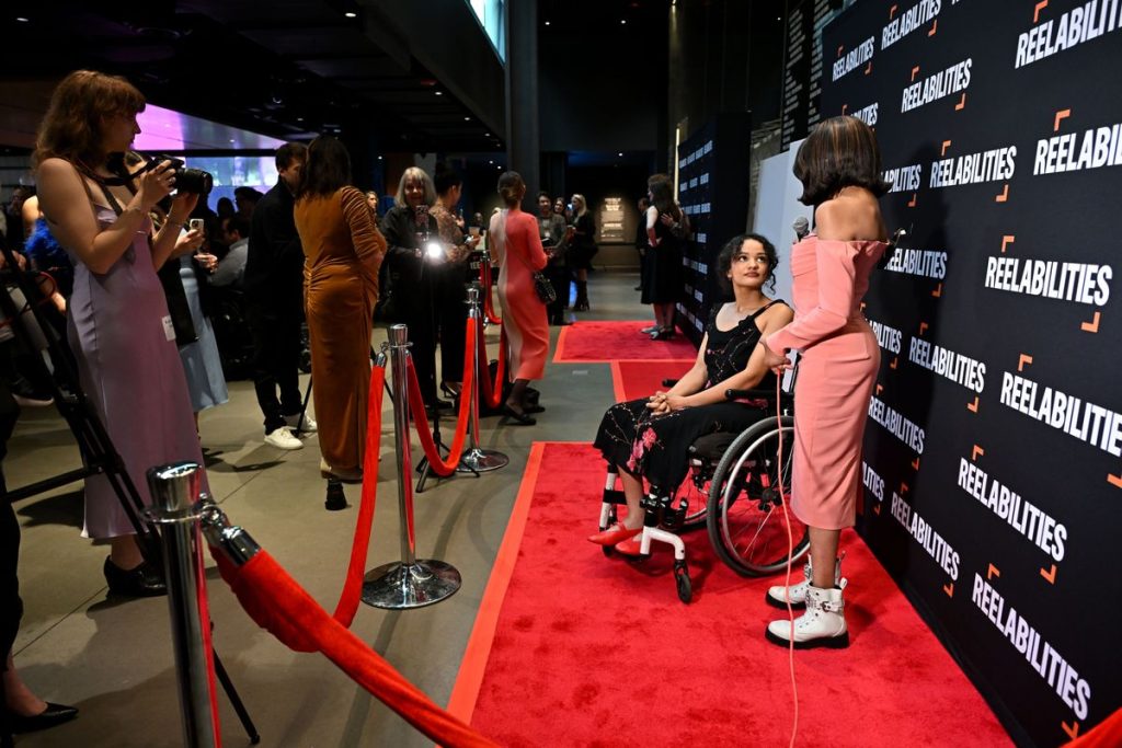 Zwei Personen in Abendgarderobe sind auf einem roten Teppich, eine nutzt einen Rollstuhl. Hinter einem roten Samtseil stehen Besuchende.