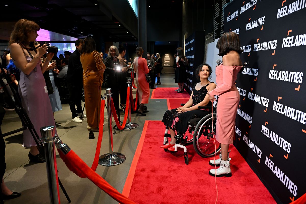 Zwei Personen in Abendgarderobe sind auf einem roten Teppich, eine nutzt einen Rollstuhl. Hinter einem roten Samtseil stehen Besuchende.
