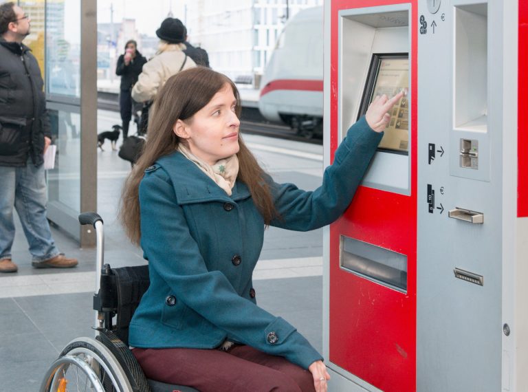 Eine Frau im Rollstuhl bedient in einem Bahnhof einen Fahrkartenautomaten der Deutschen Bahn.