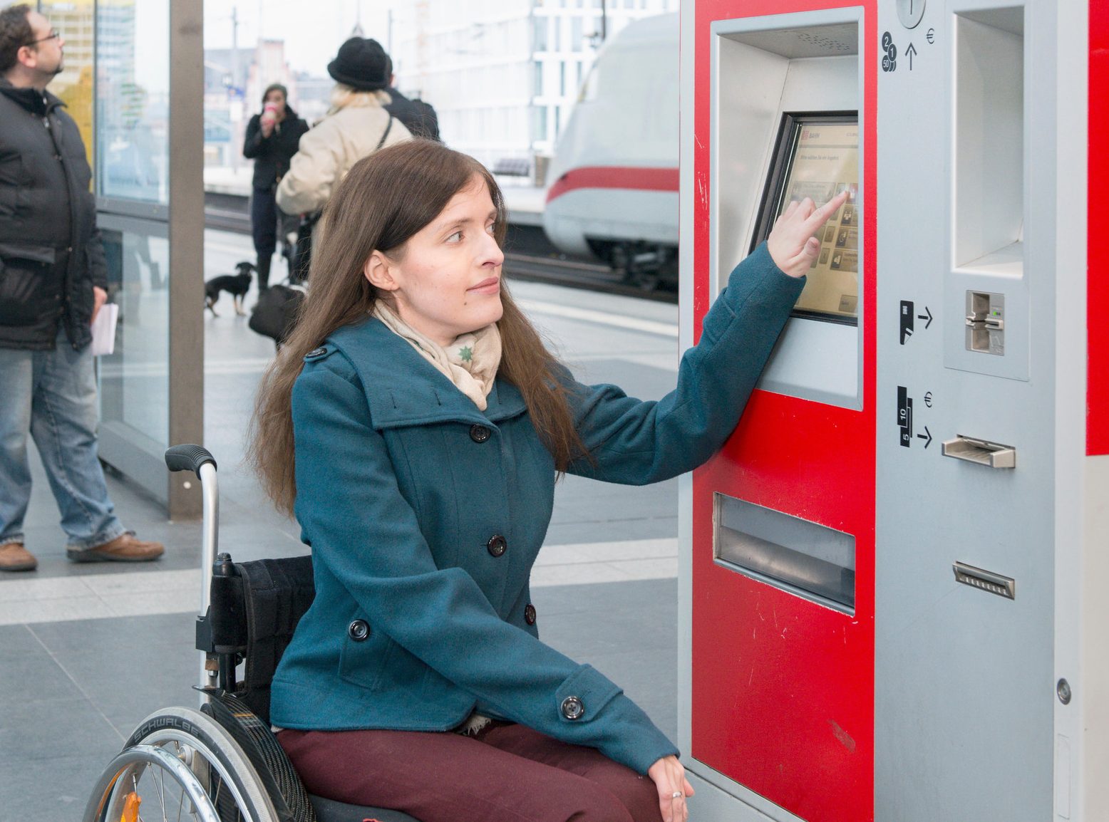 Eine Frau im Rollstuhl bedient in einem Bahnhof einen Fahrkartenautomaten der Deutschen Bahn.