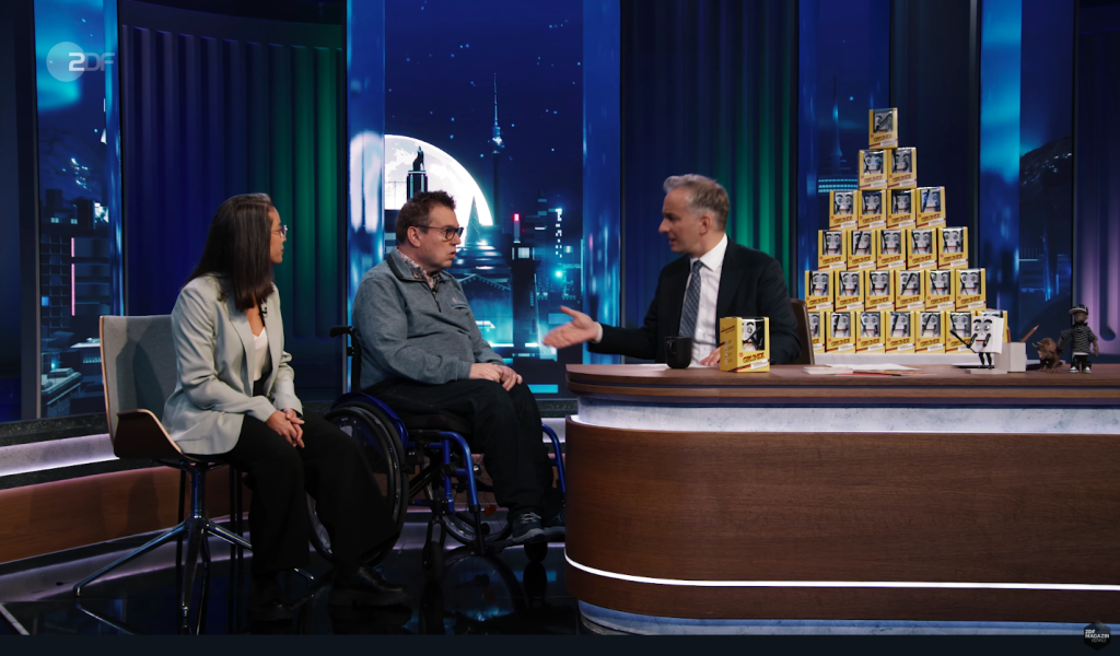 Bühne einer Fernsehshow, hinter einem Tisch sitzt Jan Böhmermann, neben ihm Jürgen Linnemann, daneben Soraia Da Costa Batista. Rechts steht eine Pyramide aus Grundi-Figuren. Im Hintergrund ist ein Nachthimmel mit Mond projeziert.