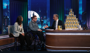 Bühne einer Fernsehshow, hinter einem Tisch sitzt Jan Böhmermann, neben ihm Jürgen Linnemann, daneben Soraia Da Costa Batista. Rechts steht eine Pyramide aus Grundi-Figuren. Im Hintergrund ist ein Nachthimmel mit Mond projeziert.