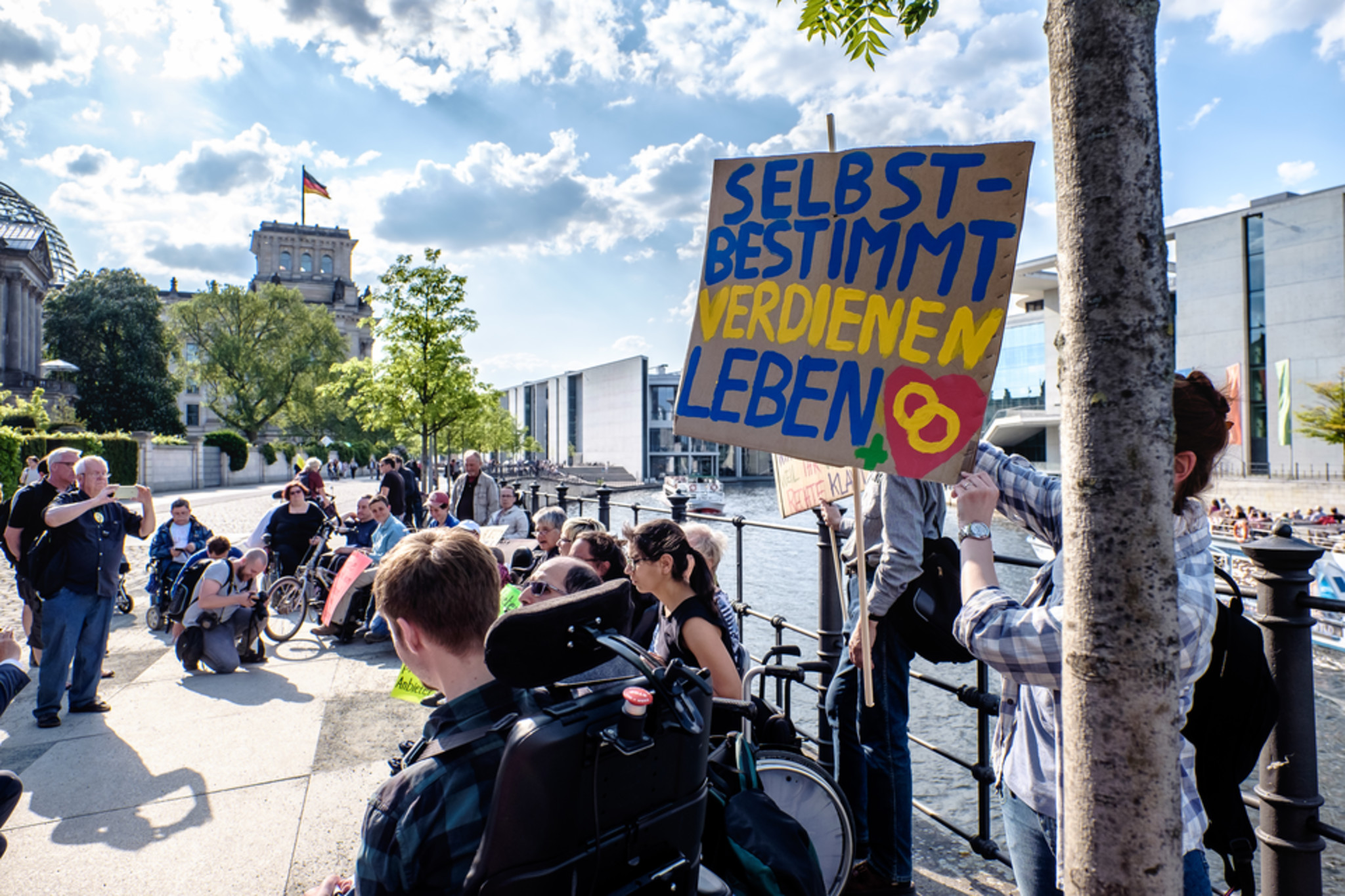Demo am Reichtagsufer, im Hintergrund der Bundestag. Die Sonne scheint, einige Menschen haben sich versammelt. Eine Person trägt ein Schild mit der Aufschrift „Selbstbestimmt Leben“