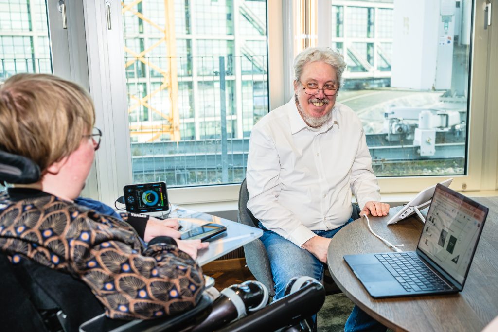 Wolfgang, ein weißer Mann mit grauen lockigen Haaren, sitzt an einem Tisch und schaut zu einer Arbeitskollegin, die im Elektrorollstuhl sitzt.