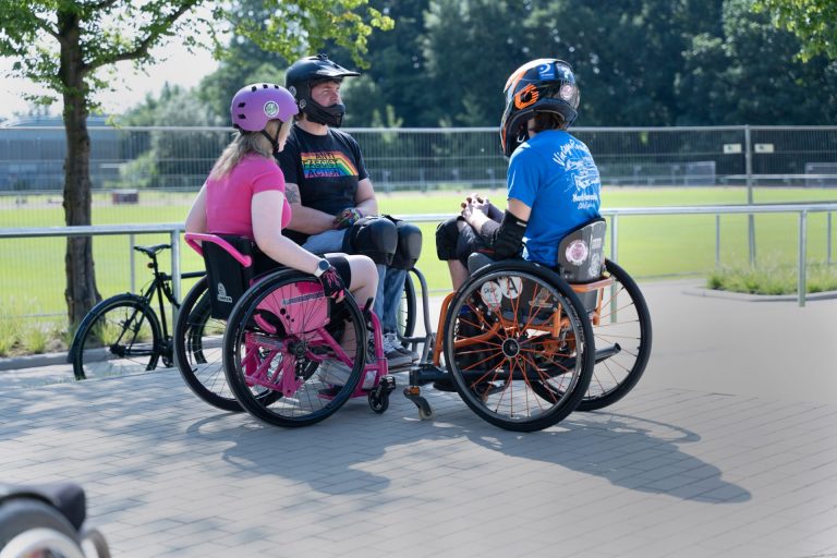 Drei Personen mit Helm im Aktiv-Rolli sitzen im Kreis auf einem Skateplatz.