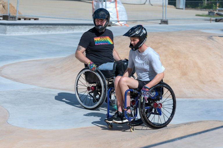 Zwei weiß gelesene Personen mit Helm im Aktivrolli auf einem Skateplatz.