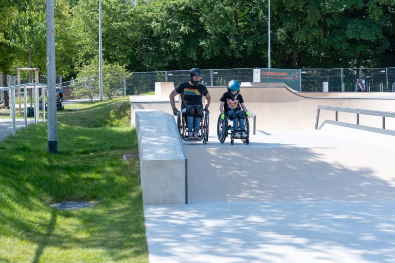 Ein Skateplatz, zwei Personen im Rolli stehen vor einem Hügel.