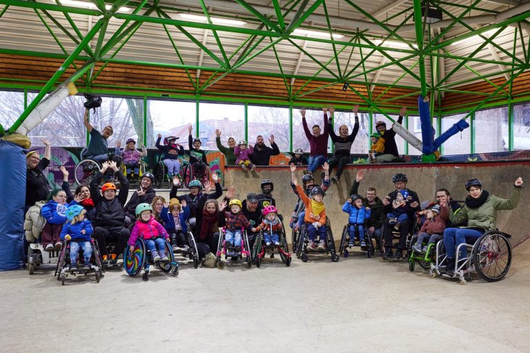 Große Gruppe an jungen und älteren Rolli-Skater*innen in einer Skatehalle.