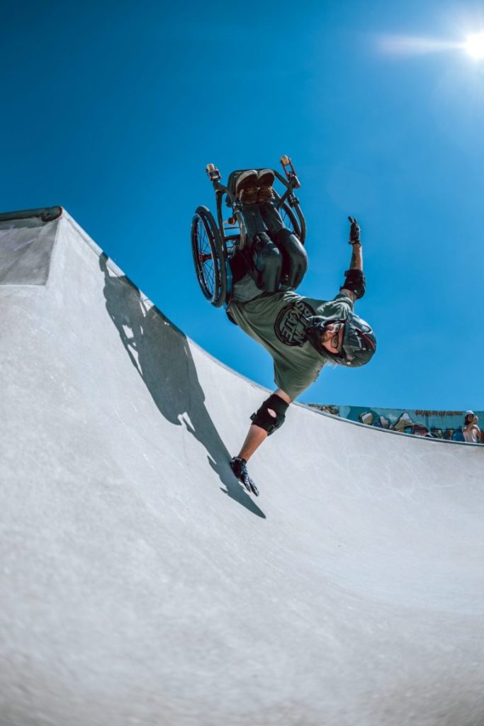 Person im Rolli in einer Half Pipe, über ihr blauer Himmel. Der Rollstuhl ist in der Luft, eine Hand ist auf dem Boden.