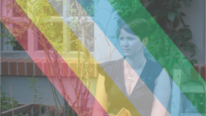 Person mit einem brauenen Kurzhaarschnitt vor einer Hauswand mit Fenstern schaut zur Seite. Sie trägt ein schwarzes V-Ausschnitt T-Shirt, mehrere Ohrringe und eine Kette. Über das Bild ist leicht transparent eine Disability Pride Flagge gelegt.