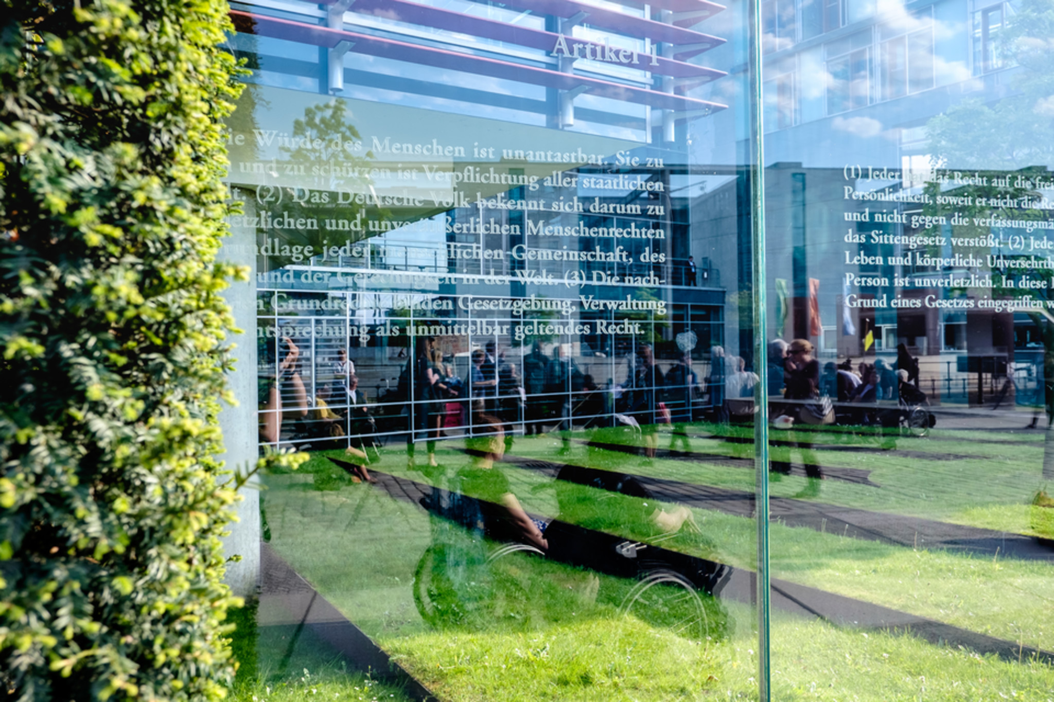 Artikel 1 des Grundgesetzes auf einer Glasscheibe, die zum Bundestagsgebäude gehört, links ist eine grüne Pflanze im Bild, es spiegeln sich leicht einige Personen im Rollstuhl.