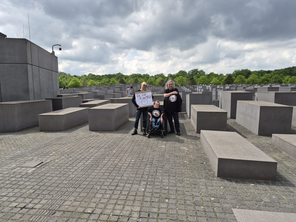 3 Personen im schwazen Shirt vor dem Holocaust Denkmal in Berlin. Eine Person trägt ein Schild: 5 vor 1933. 1 Person nutzt einen Rollstuhl.