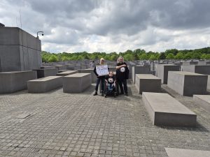3 Personen im schwazen Shirt vor dem Holocaust Denkmal in Berlin. Eine Person trägt ein Schild: 5 vor 1933. 1 Person nutzt einen Rollstuhl.