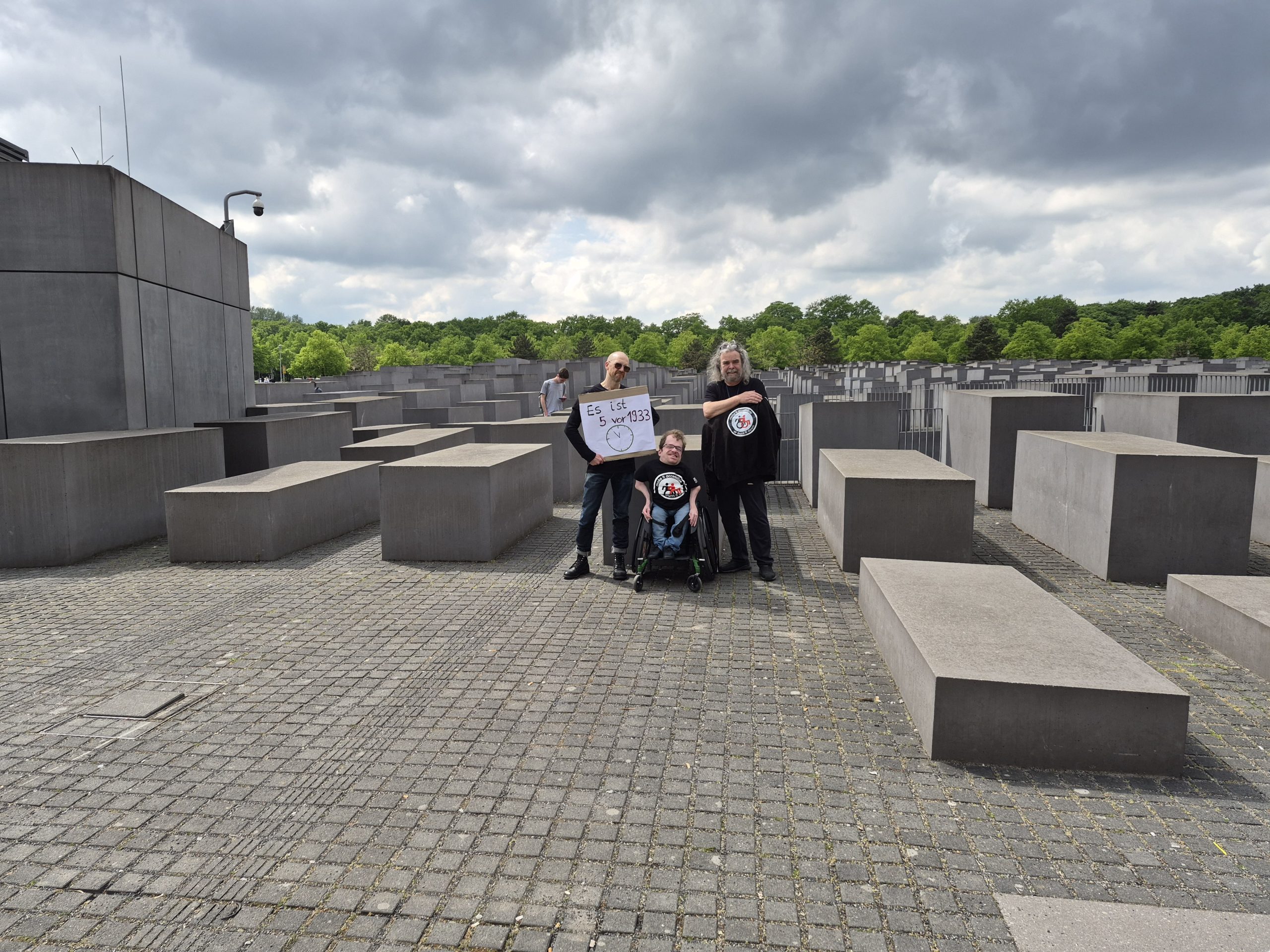 3 Personen im schwazen Shirt vor dem Holocaust Denkmal in Berlin. Eine Person trägt ein Schild: 5 vor 1933. 1 Person nutzt einen Rollstuhl.