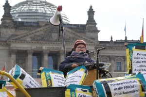 Mehrere Baueimer und Zementsäcke sind vor dem Bundestag aufgebaut, eine Person im Rolli spricht in ein Mikrofon.