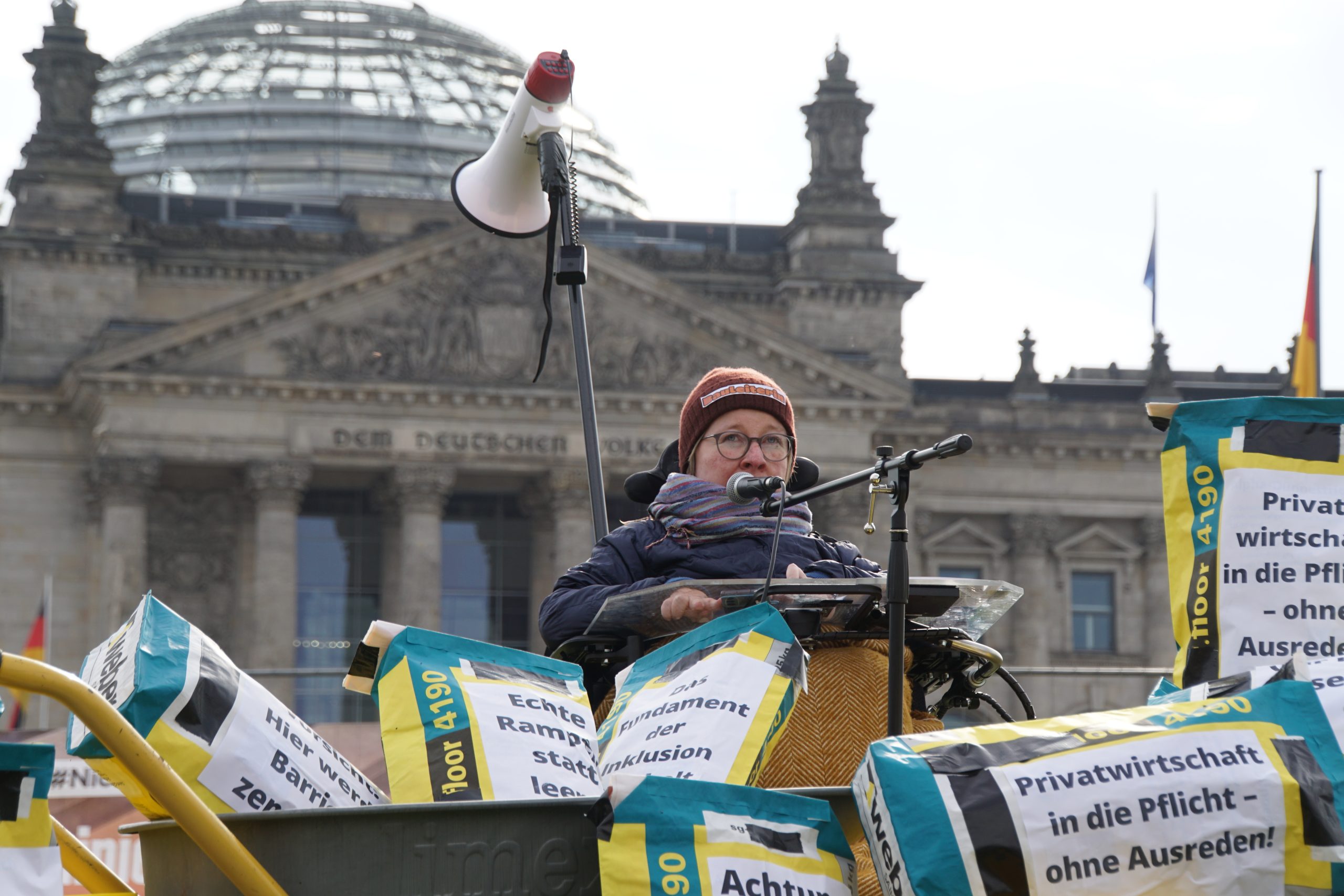 Mehrere Baueimer und Zementsäcke sind vor dem Bundestag aufgebaut, eine Person im Rolli spricht in ein Mikrofon.