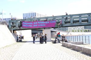 An einer Brücke im Regierungsviertel in Berlin hängt das Plakat „Krüppel die kämpfen sind Krüppel die leben.“ Zwei Personen im Rollstuhl haben sich von der Brücke abgeseilt. Eine Gruppe Polizist*innen steht davor.