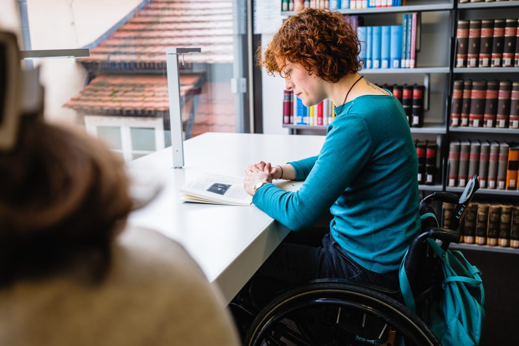 Person mit halblangen, roten Locken sitzt an einem Tisch in der Bibliothek, sie nutzt einen Rollstuhl. Im Vordergrund ist eine zweite Person angeschnitten.