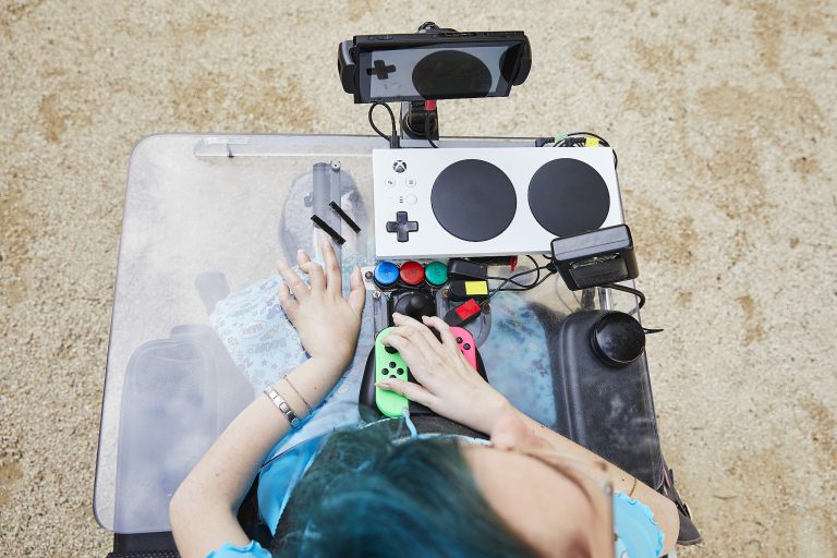 Blcik von oben auf eine Person, die im Rollstuhl sitzt. Vor sich hat sie verschiedene Gaming-Controller.