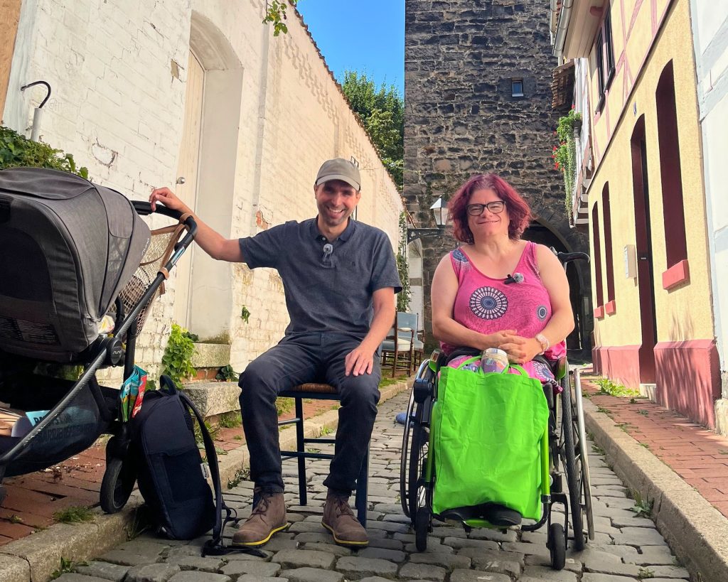 Zwei Personen sitzen in einer schmalen Gasse, die linke trägt dunkle Kleidung und hat eine Hand an einem Kinderwagen, die rechte trägt einen knalligen grünen Rock und ein pinkes Shirt und nutzt einen Rollstuhl. Beide lächeln in die Kamera.