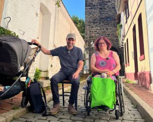 Zwei Personen sitzen in einer schmalen Gasse, die linke trägt dunkle Kleidung und hat eine Hand an einem Kinderwagen, die rechte trägt einen knalligen grünen Rock und ein pinkes Shirt und nutzt einen Rollstuhl. Beide lächeln in die Kamera.