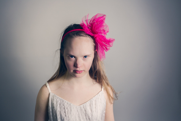 Weiß positionierte Person mit langen, brauen Haaren und pinker Federboa auf dem Kopf schaut ernst von schräg unten in die Kamera. Sie trägt ein weißes Trägerkleid.