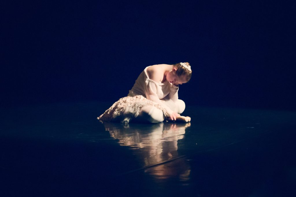 Auf einer schwarzen Theaterbühne sitzt eine junge Frau im Scheinwerferlicht. Sie hat ihren Oberkörper nach vorne gebeugt und schaut auf den Boden.