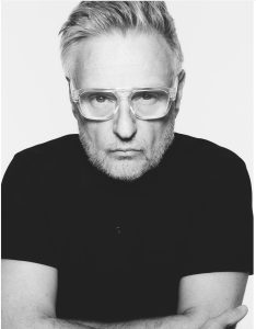 Schwarz-weiß Porträt einer weiß positionierten Person mit kurzen, grauen Haaren, einer großen, durchsichtig umrandeten Brille und einem schwarzen Shirt. Die Person schaut ernst in die Kamera.