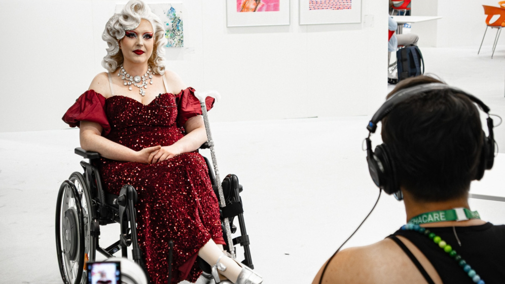 Person mit schulterfreiem, rot-glitzerndem Ballkleid und silbernen High-Heels, grauer Dauerwelle und glitzerndem Diadem sitzt in einem Rollstuhl, dahinter eine weiße Wand mit Kunst, ihr gegenüber eine Person mit Headset und Kamera.