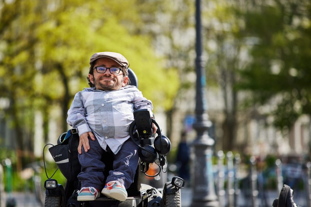 Person im E-Rollstuhl mit Schirmmütze, karriertem Hemd, Brille und Bart auf einem Gehweg, im Hintergrund Bäume und eine Laterne.