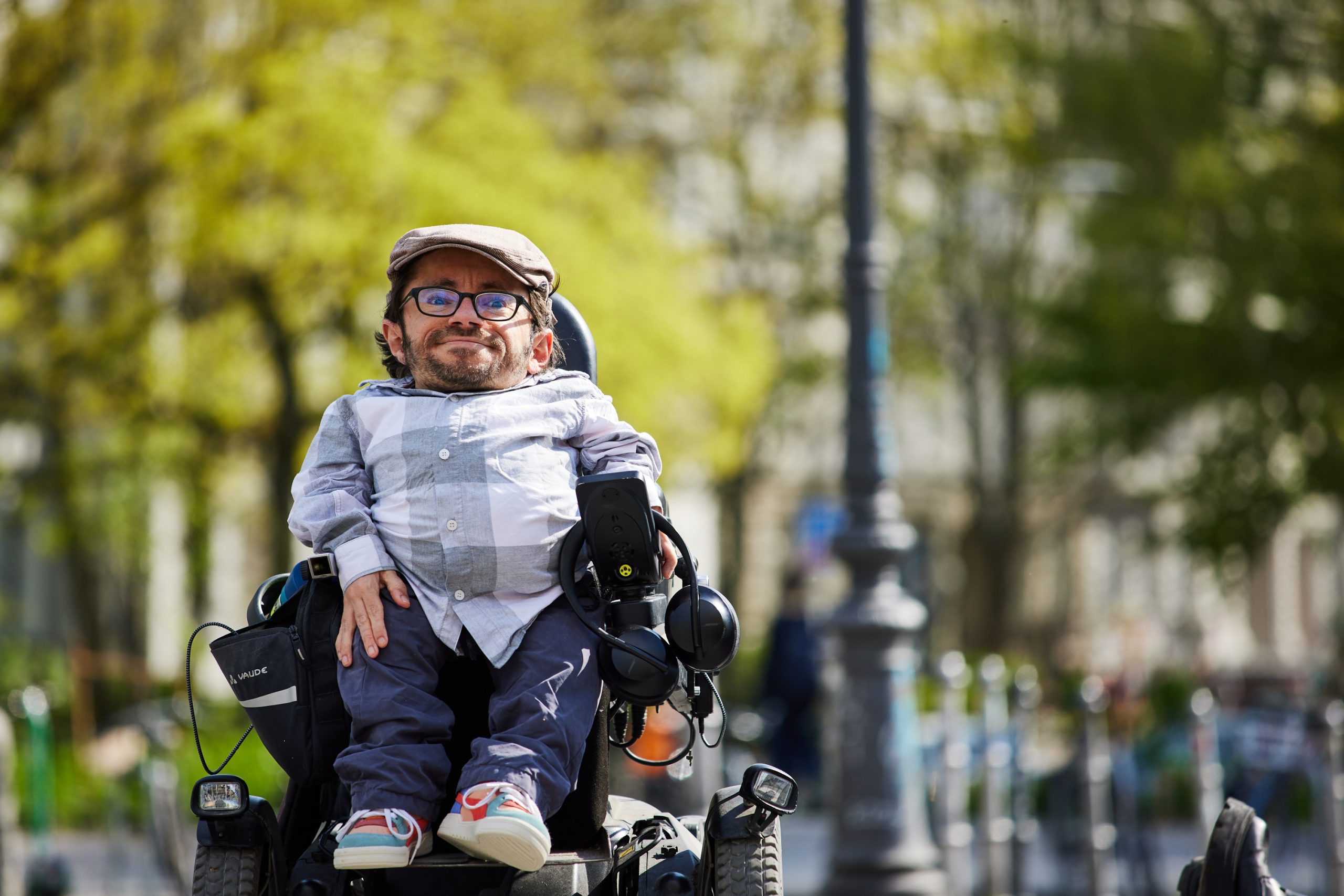 Person im E-Rollstuhl mit Schirmmütze, karriertem Hemd, Brille und Bart auf einem Gehweg, im Hintergrund Bäume und eine Laterne.